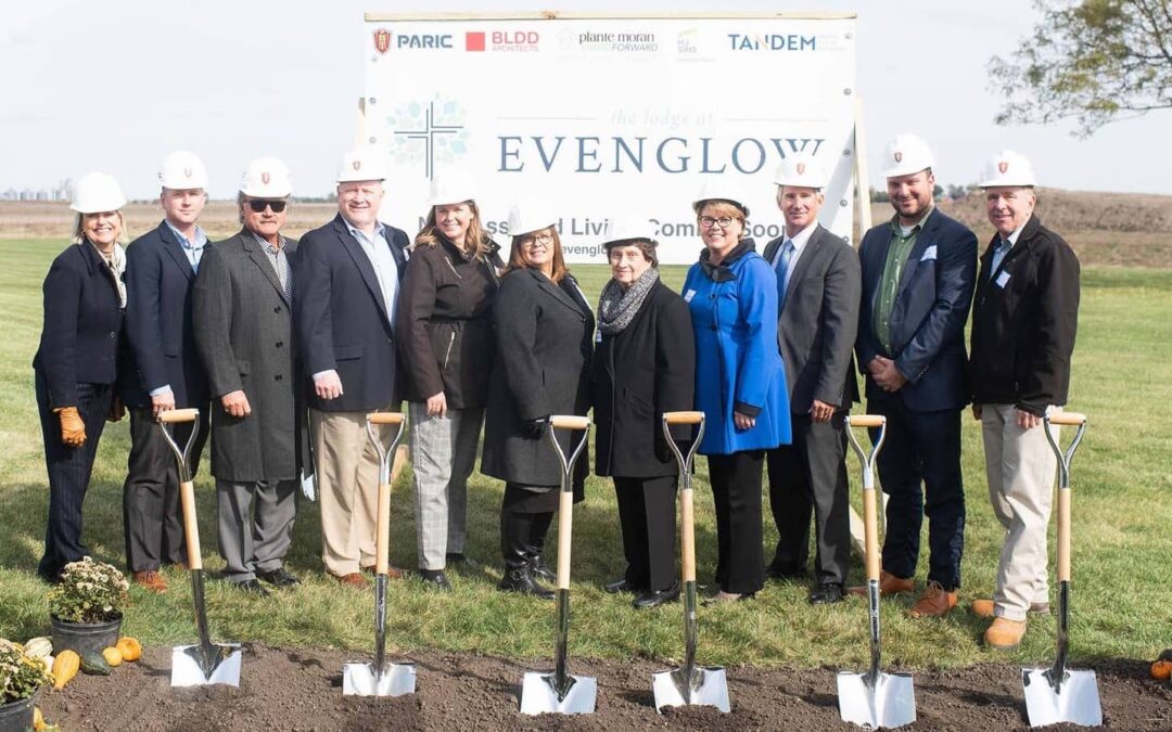 Group of people dressed in business attire and hardhats standing behind silver shovels with a sign in the background that says "Evenglow"